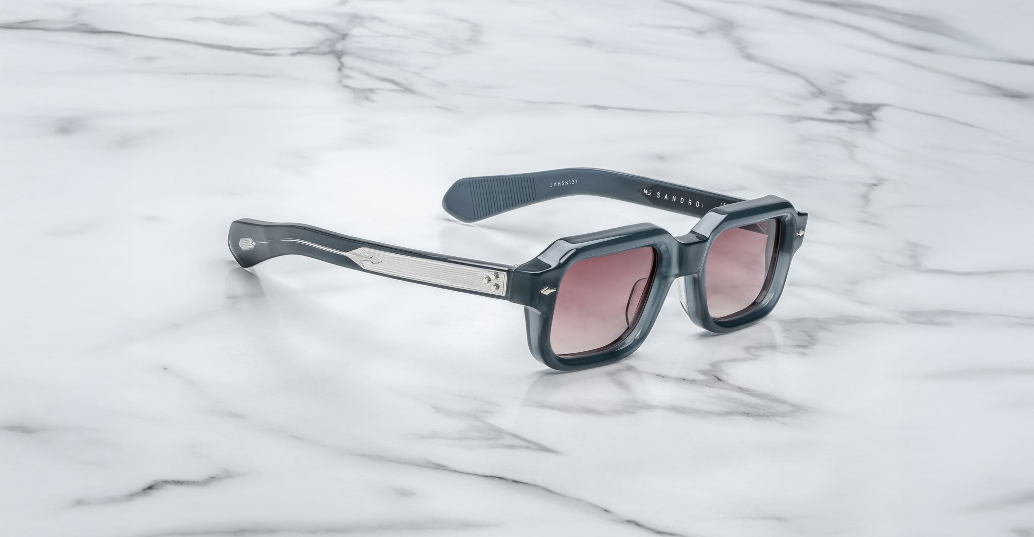 A pair of Jacques Marie Mage Sandro sunglasses with black square frames, metallic arm details, and pink-tinted lenses rests on a white marble surface.