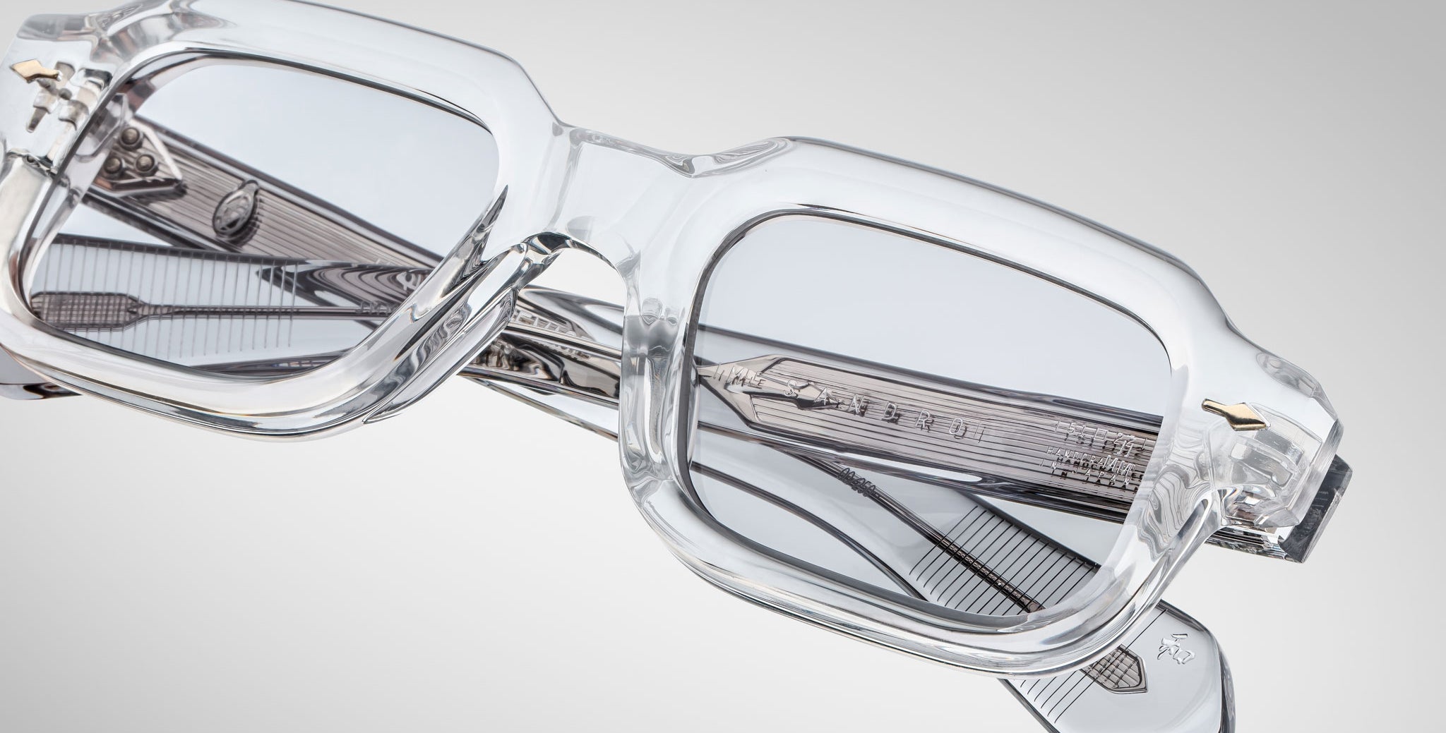 Close-up of Jacques Marie Mage Sandro clear rectangular eyeglass frames with gray-tinted lenses and intricate arm detailing, set against a light gray background.