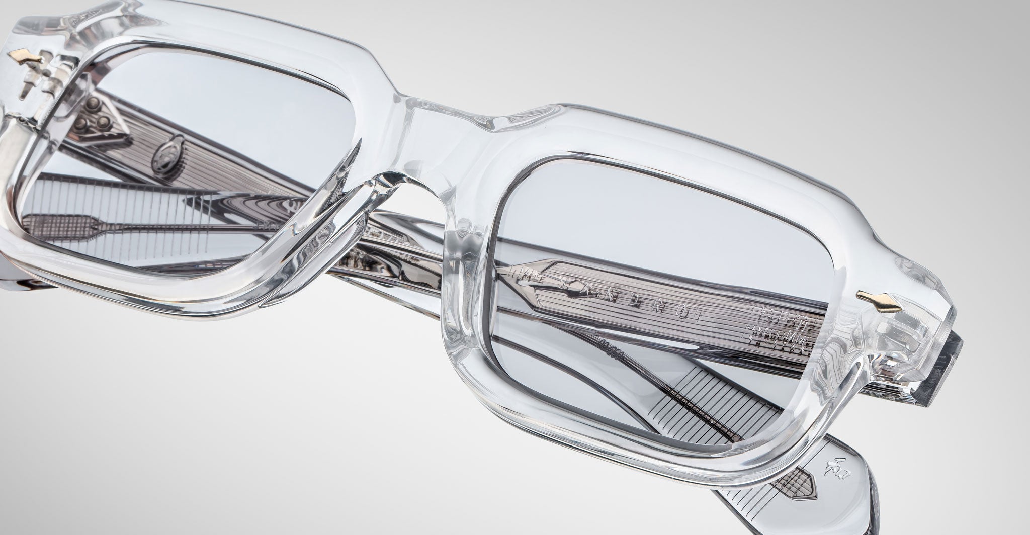 Close-up of Jacques Marie Mage Sandro clear rectangular eyeglass frames with gray-tinted lenses and intricate arm detailing, set against a light gray background.