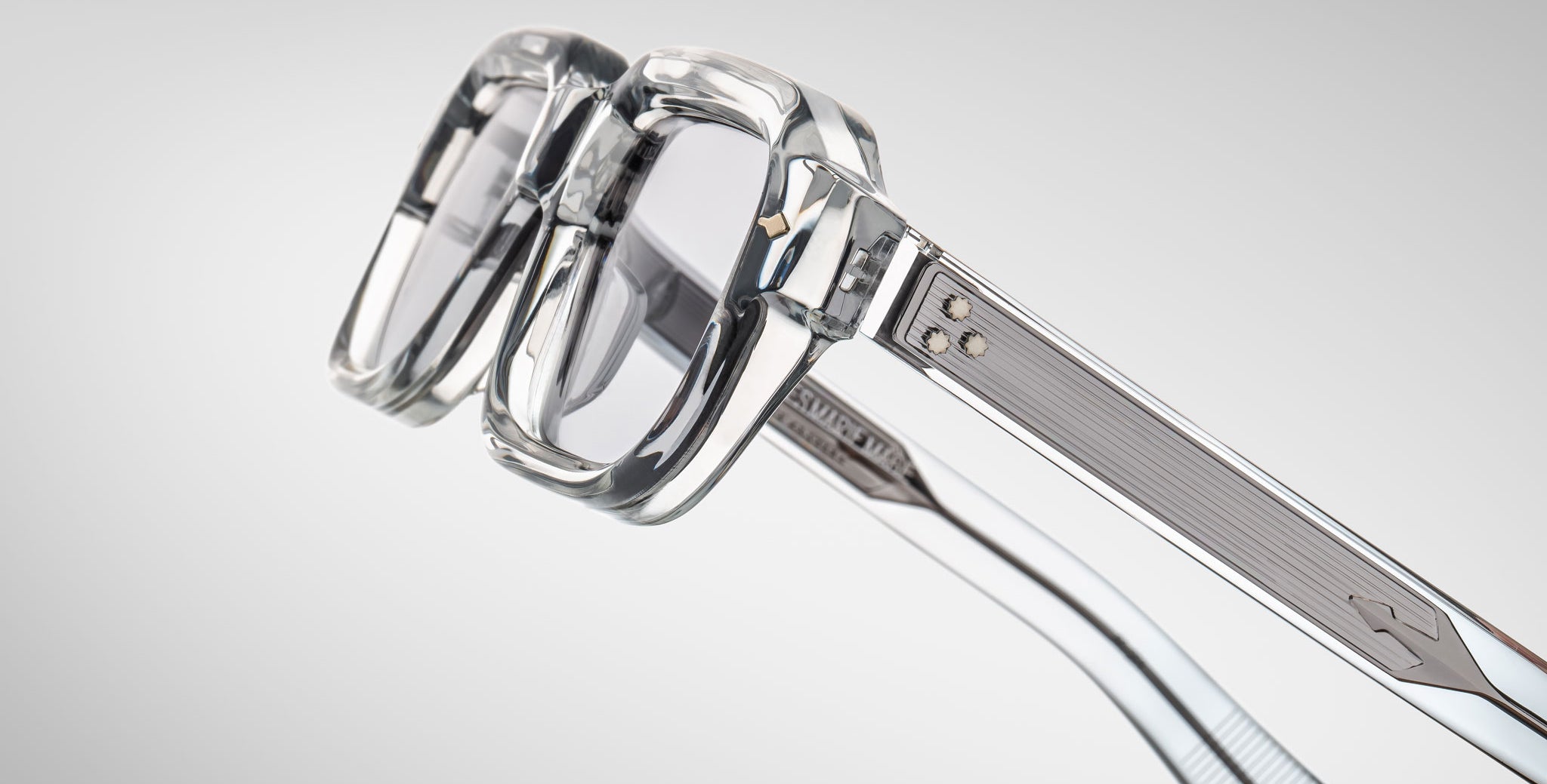 Close-up view of Jacques Marie Mage Sandro eyeglasses featuring clear, thick rims, transparent lenses, and metallic accents on the arms, set against a light gray background.