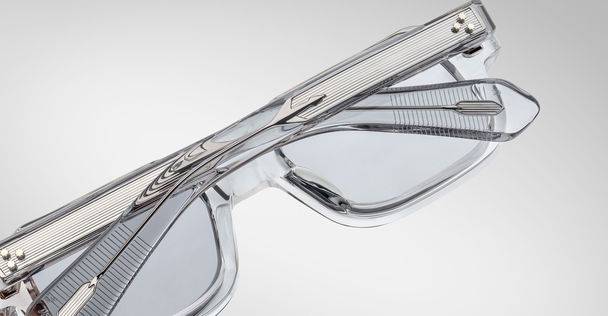 A close-up of Jacques Marie Mage Sandro eyeglasses with transparent frames and silver metallic accents, displayed from above with the temples crossed over the lenses on a plain light background.
