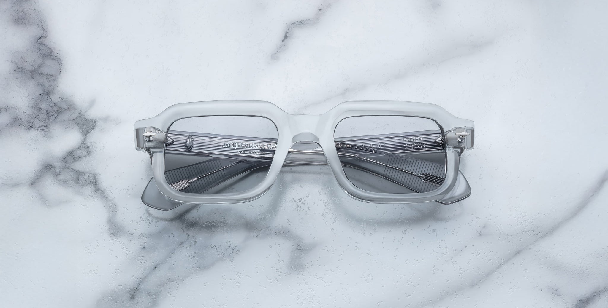 The Jacques Marie Mage Sandro eyeglasses, featuring clear rectangular frames and grey-tinted lenses, rest on a white marble surface with grey veining.