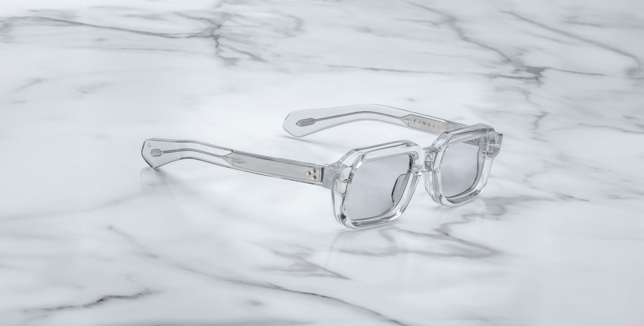 A pair of Jacques Marie Mage Sandro clear, square-framed sunglasses with silver-tinted lenses is displayed on a white marble surface.