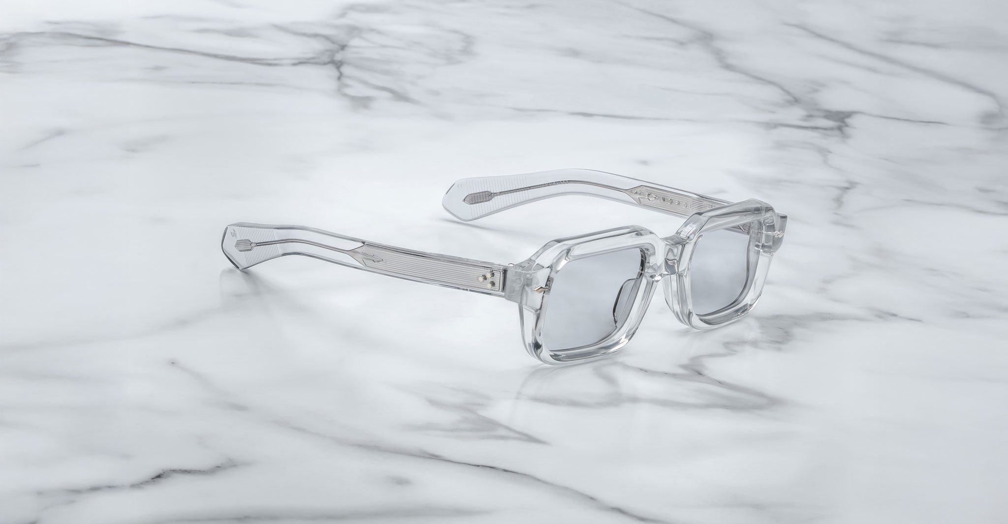 A pair of Jacques Marie Mage Sandro clear, square-framed sunglasses with silver-tinted lenses is displayed on a white marble surface.