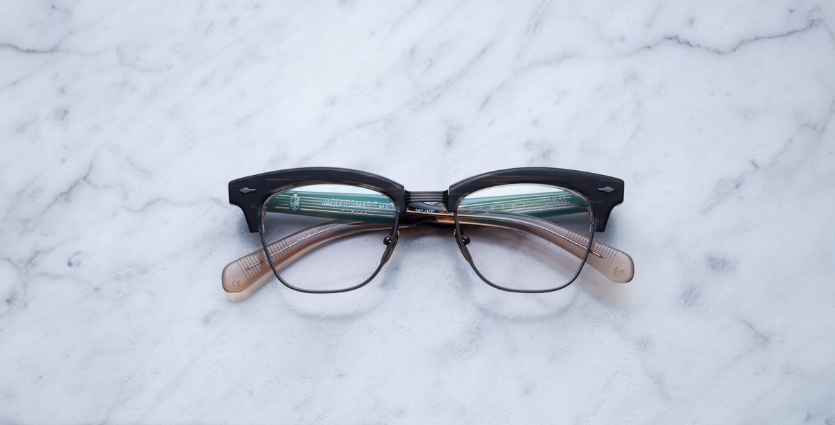 A pair of Jacques Marie Mage Sartre glasses with black and translucent brown clubmaster optical frames rests on a white marble surface.