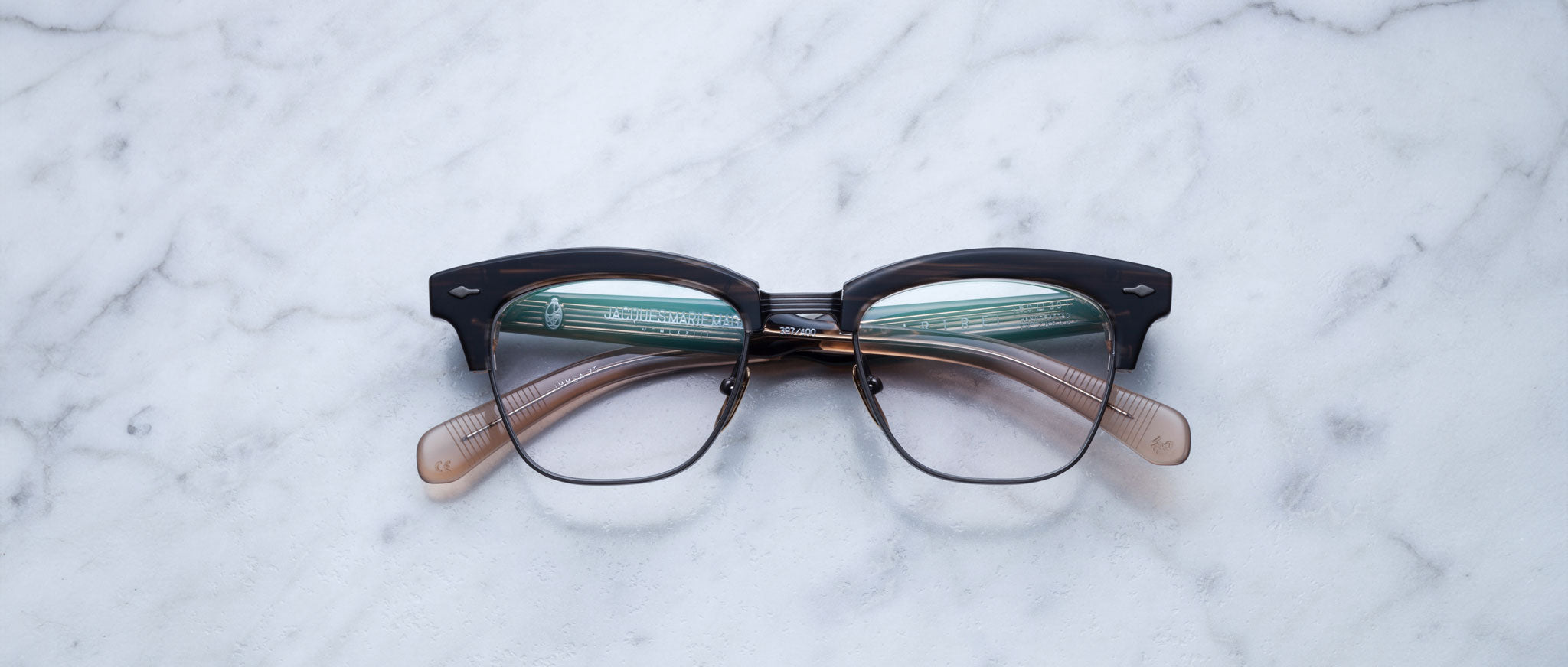 A pair of Jacques Marie Mage Sartre glasses with black and translucent brown clubmaster optical frames rests on a white marble surface.