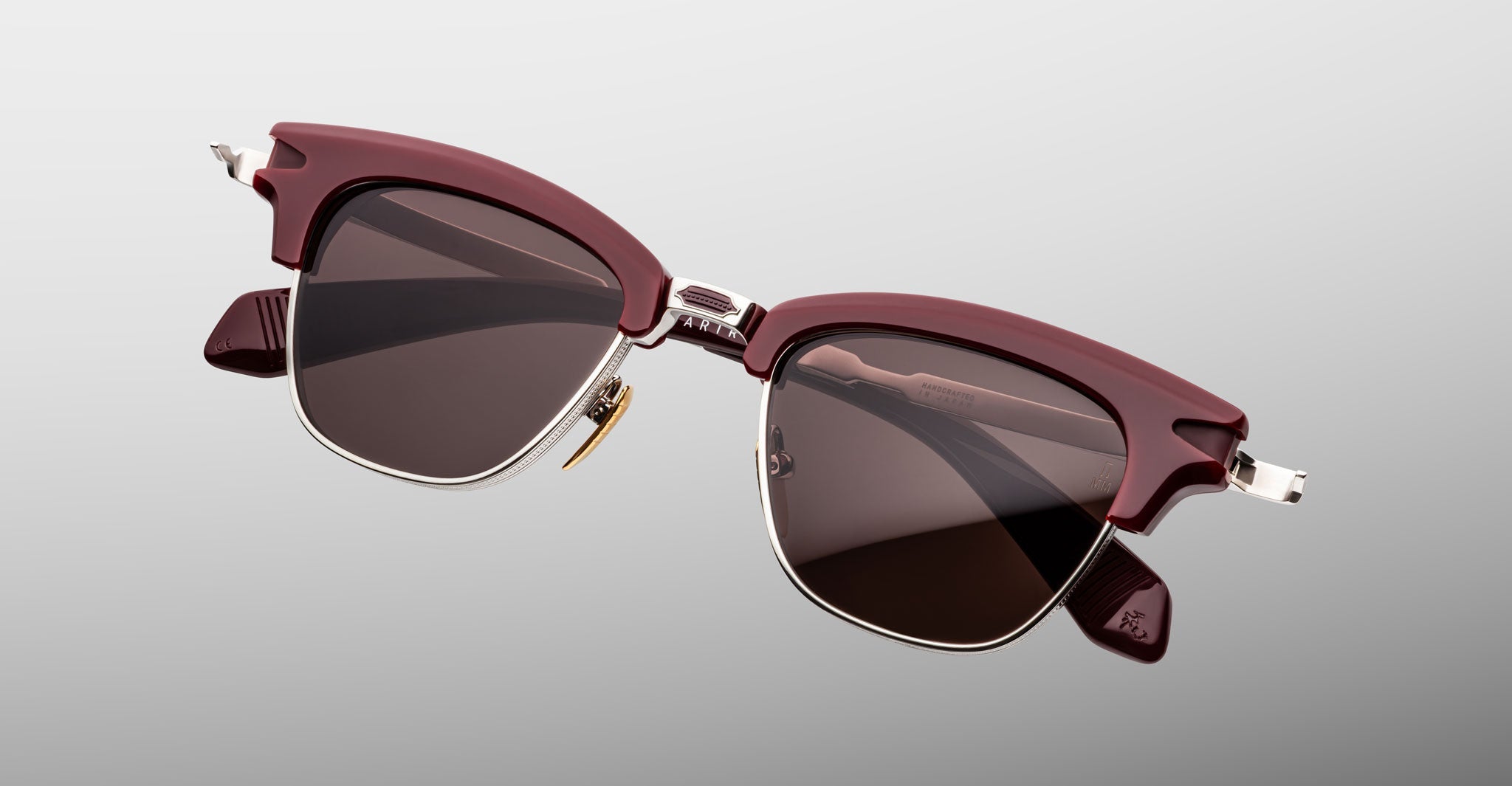 A pair of Jacques Marie Mage Sartre glasses with dark lenses and a maroon and silver clubmaster frame, displayed against a gradient gray background.