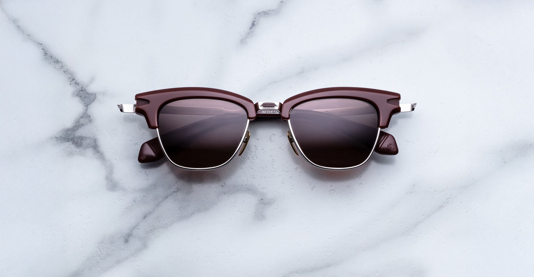 Jacques Marie Mage Sartre sunglasses in brown and silver with dark lenses rest on a white marble surface.