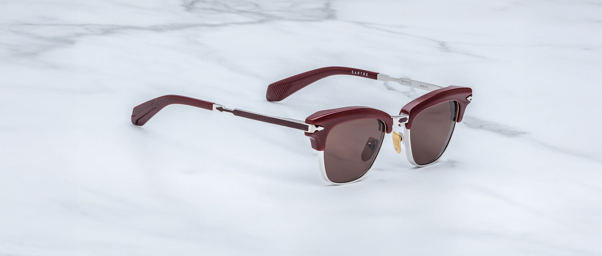 A pair of Jacques Marie Mage Sartre glasses with maroon frames, silver accents, and dark lenses rests on a white marble surface.