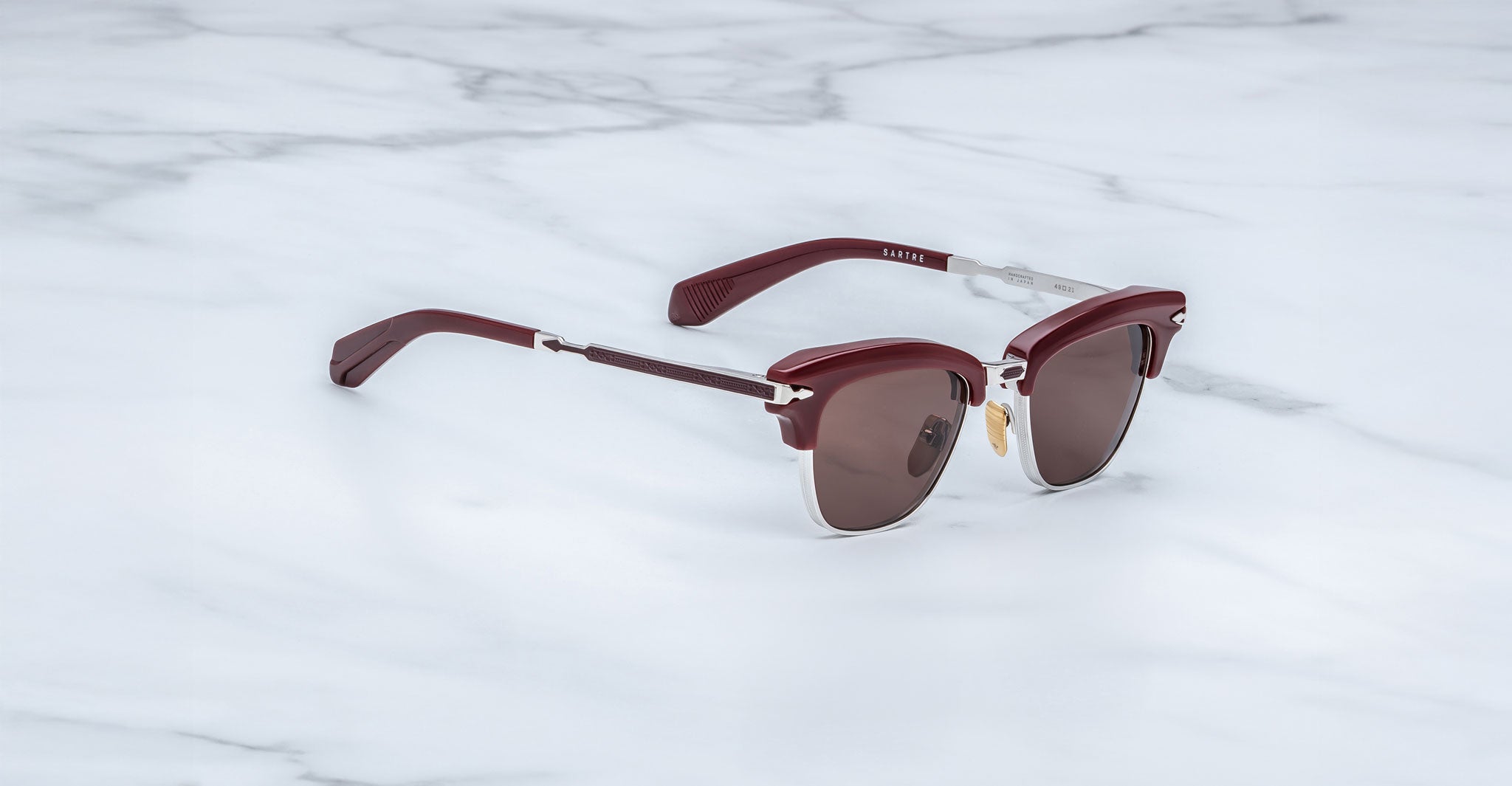 A pair of Jacques Marie Mage Sartre glasses with maroon frames, silver accents, and dark lenses rests on a white marble surface.