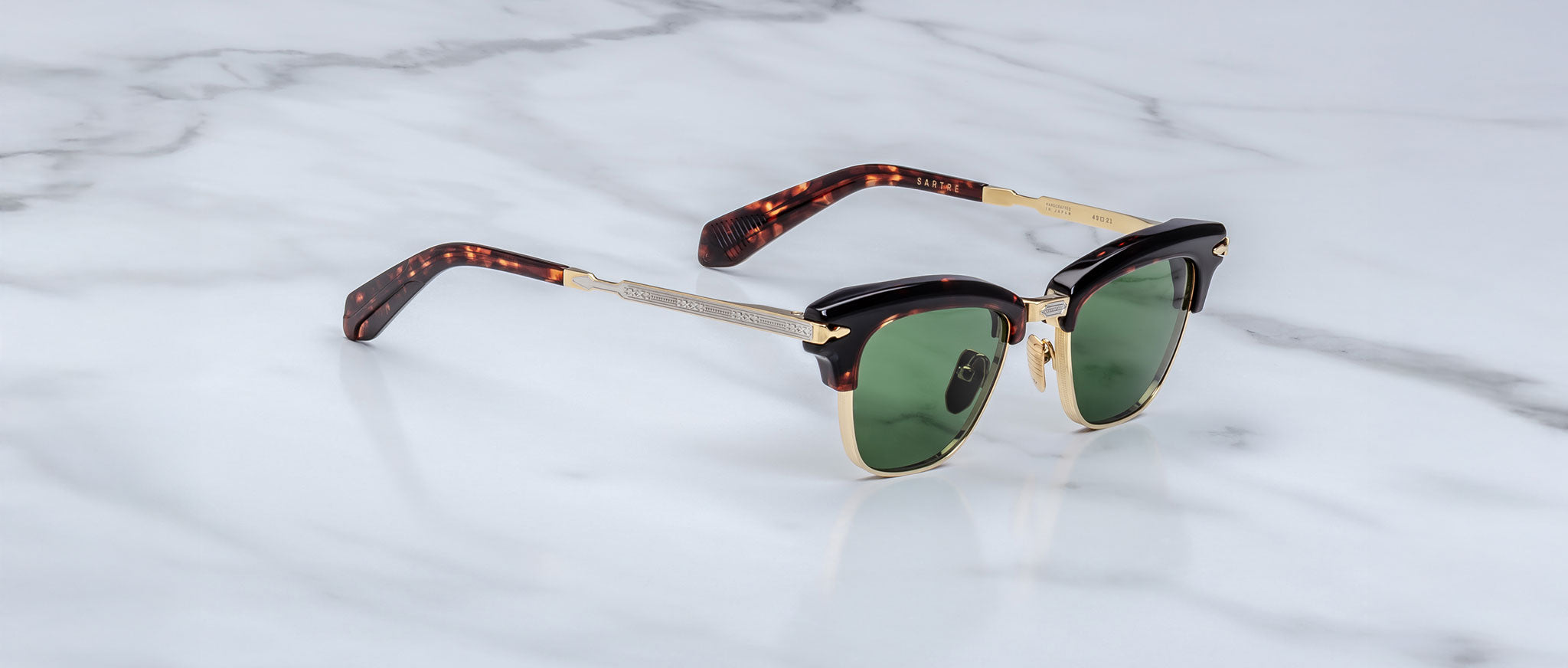 A pair of Jacques Marie Mage Sartre glasses with gold metal frames, tortoiseshell-patterned arms, and green lenses sits on a white marble surface.
