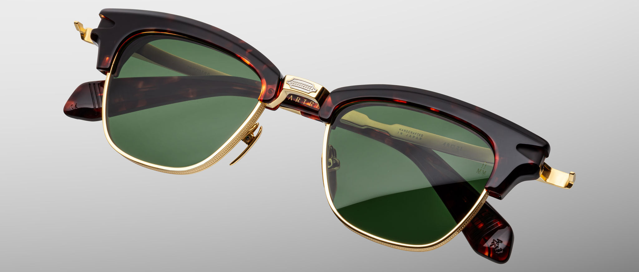 The Jacques Marie Mage Sartre sunglasses by Jacques Marie Mage feature tortoiseshell and gold frames with green lenses, inspired by classic clubmaster design, displayed on a neutral gray background.