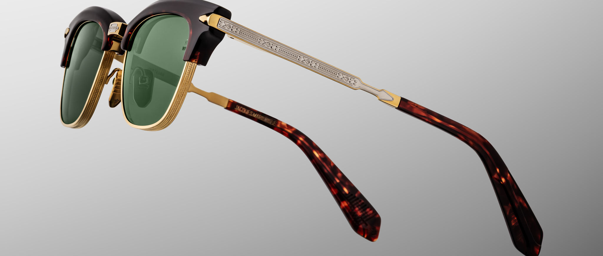 Jacques Marie Mage Sartre glasses with green lenses, gold metal accents, engraved silver arms, and tortoiseshell-patterned temple tips are shown on a gray background.