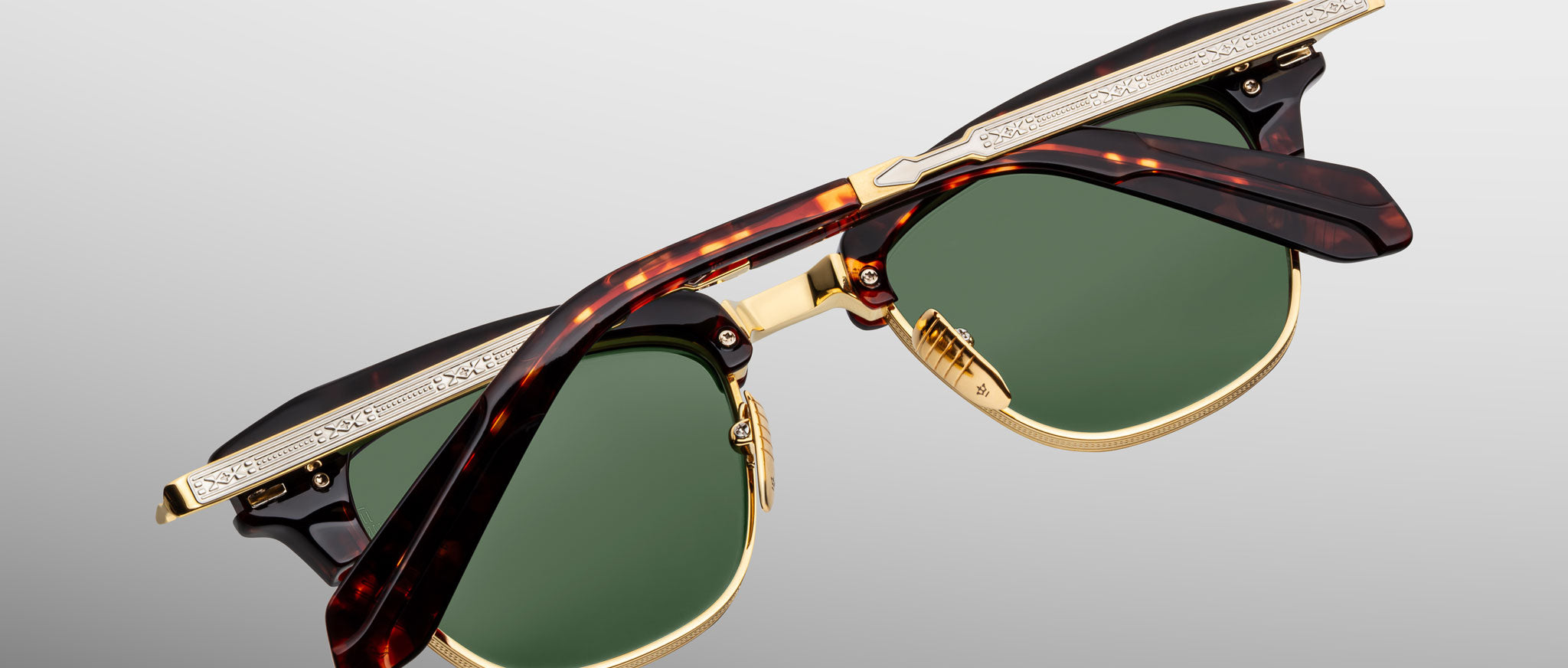 A pair of Jacques Marie Mage Sartre glasses with gold frames, green lenses, and tortoiseshell-patterned arms is displayed folded against a light gray background.