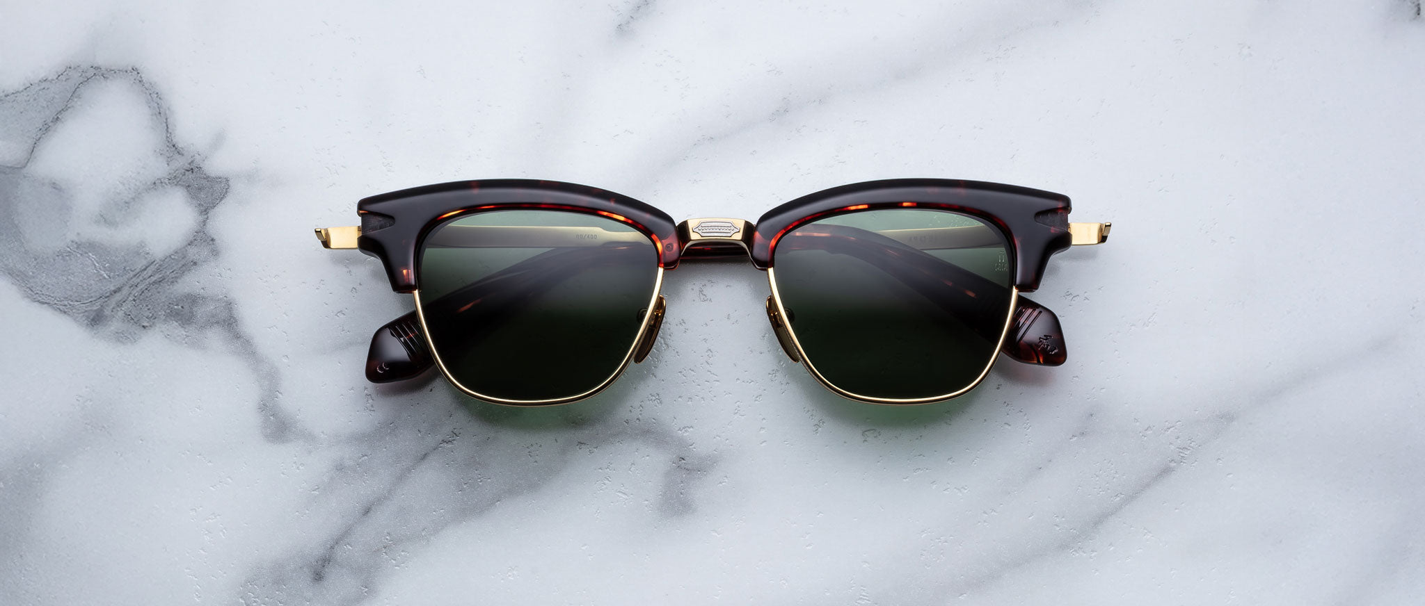 A pair of Jacques Marie Mage Sartre glasses in brown and gold with dark lenses rests on a white marble surface.