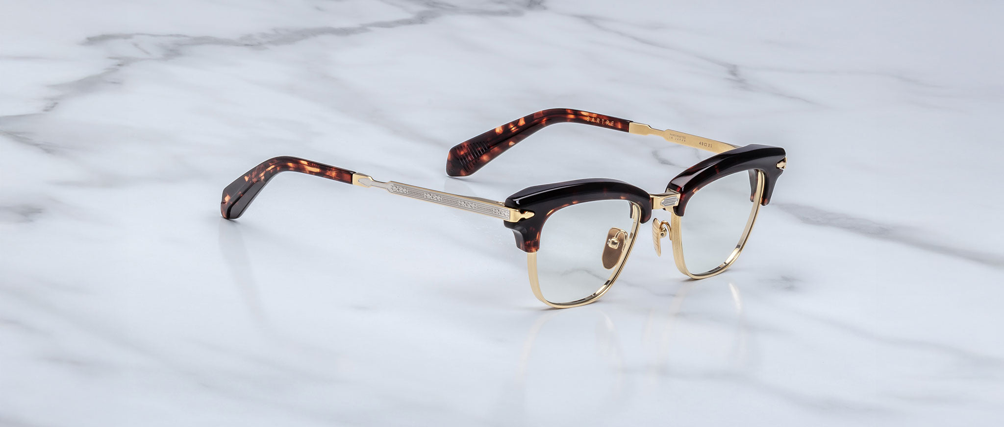 The Jacques Marie Mage Sartre glasses by Jacques Marie Mage feature gold rims, black upper frames, and tortoiseshell-patterned arms in a classic clubmaster style, shown on a white marble surface.