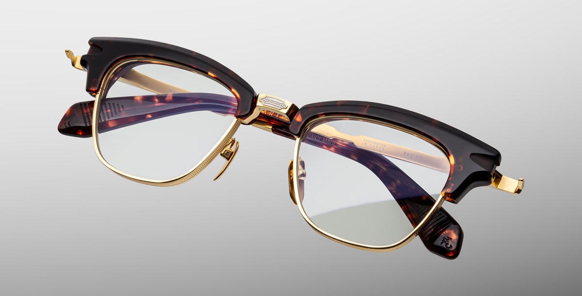 The Jacques Marie Mage Sartre glasses feature tortoiseshell-patterned clubmaster optical frames with gold accents and clear lenses, shown on a light gray background.