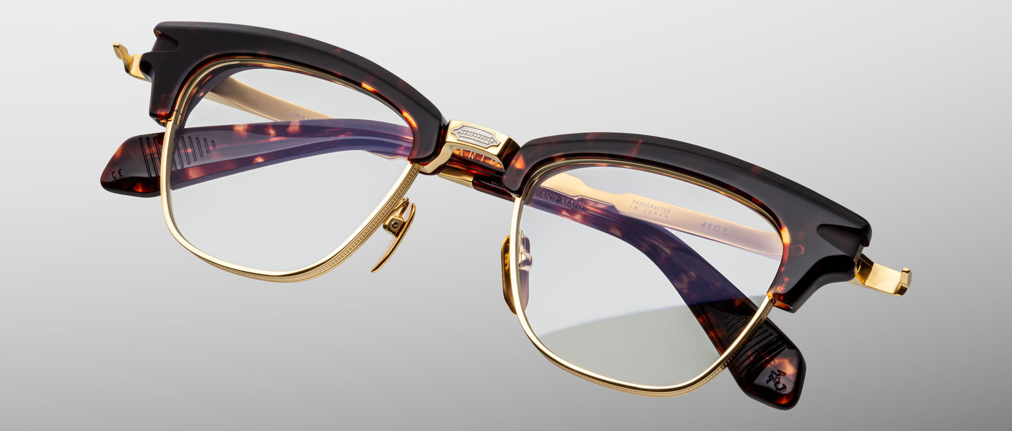 The Jacques Marie Mage Sartre glasses feature tortoiseshell-patterned clubmaster optical frames with gold accents and clear lenses, shown on a light gray background.