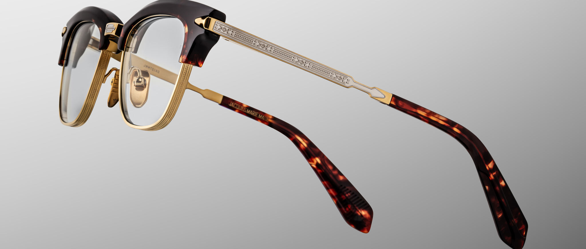 The Jacques Marie Mage Sartre glasses feature gold metal frames, a tortoiseshell-patterned browline and temple tips, and clear nose pads, displayed against a neutral background.