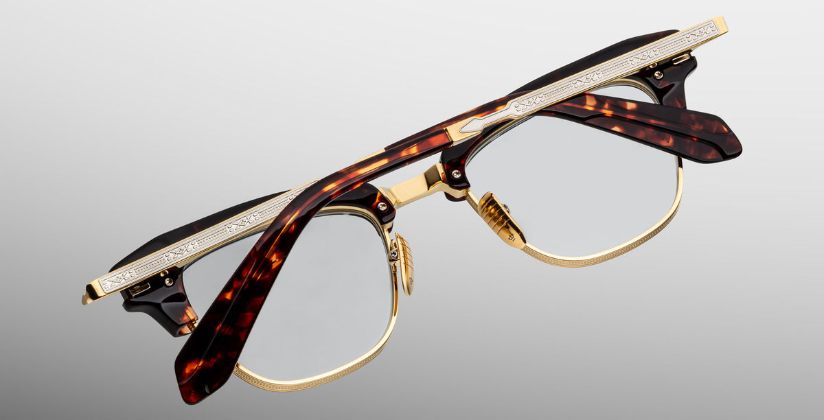 A pair of Jacques Marie Mage Sartre glasses with gold metal rims and tortoiseshell-patterned frames, engraved temples, by Jacques Marie Mage, photographed on a light gray background.
