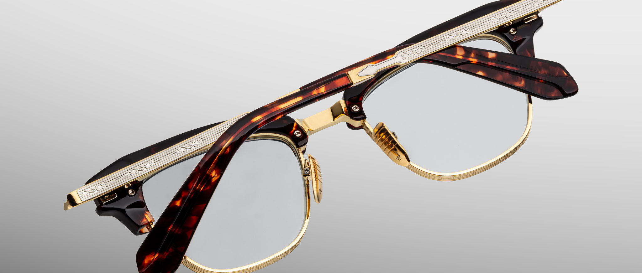 A pair of Jacques Marie Mage Sartre glasses with gold metal rims and tortoiseshell-patterned frames, engraved temples, by Jacques Marie Mage, photographed on a light gray background.