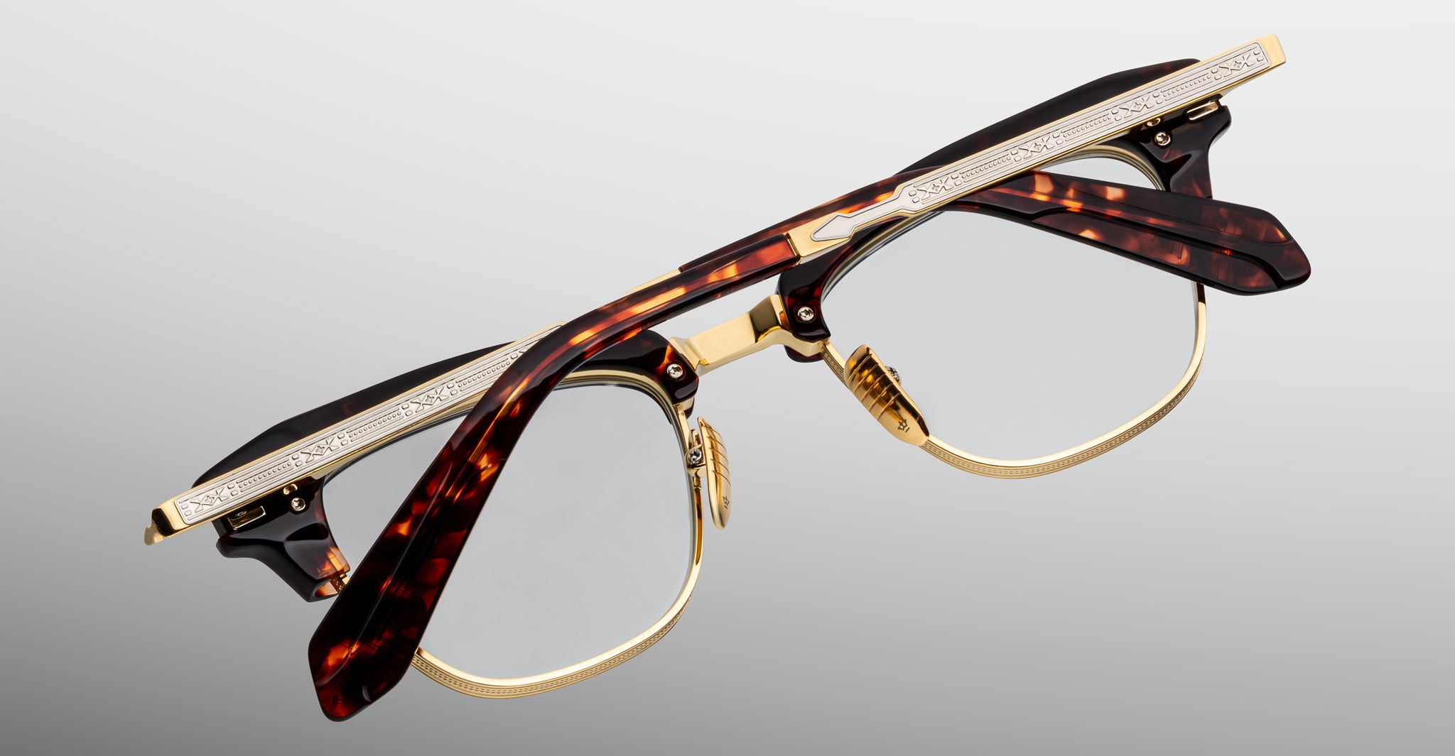 A pair of Jacques Marie Mage Sartre glasses with gold metal rims and tortoiseshell-patterned frames, engraved temples, by Jacques Marie Mage, photographed on a light gray background.