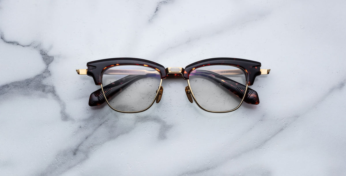 A pair of Jacques Marie Mage Sartre glasses with dark, thick frames and gold accents rests on a white marble surface.