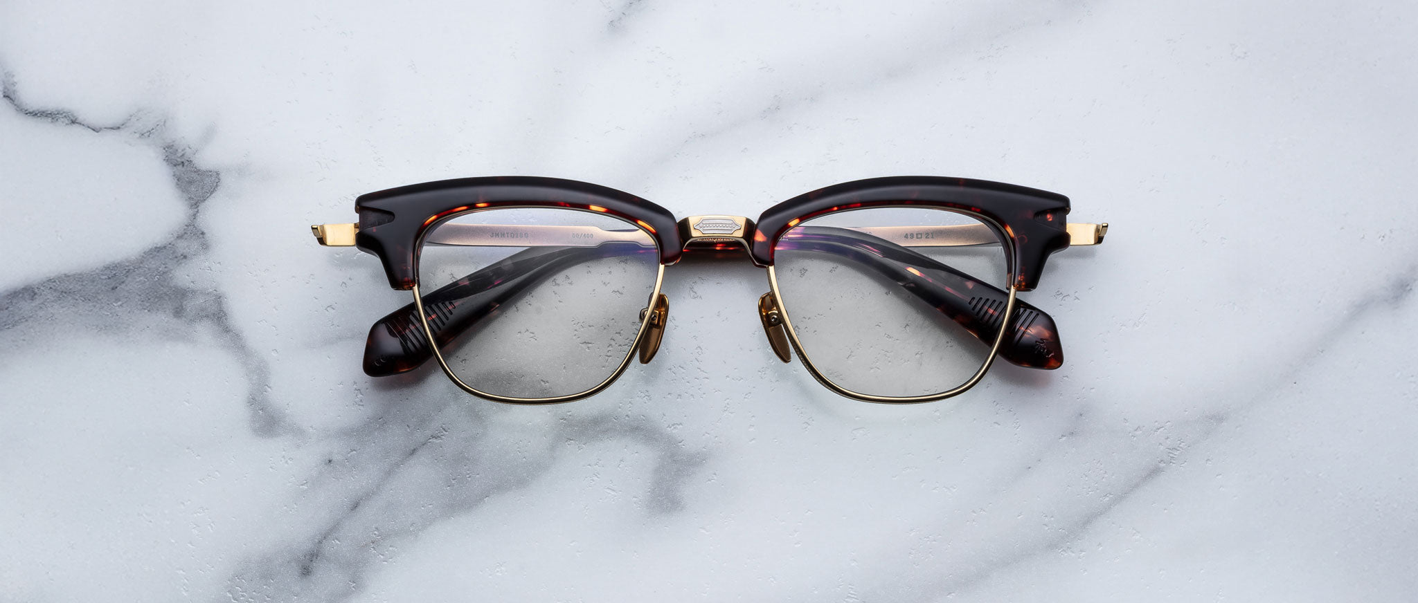 A pair of Jacques Marie Mage Sartre glasses with dark, thick frames and gold accents rests on a white marble surface.