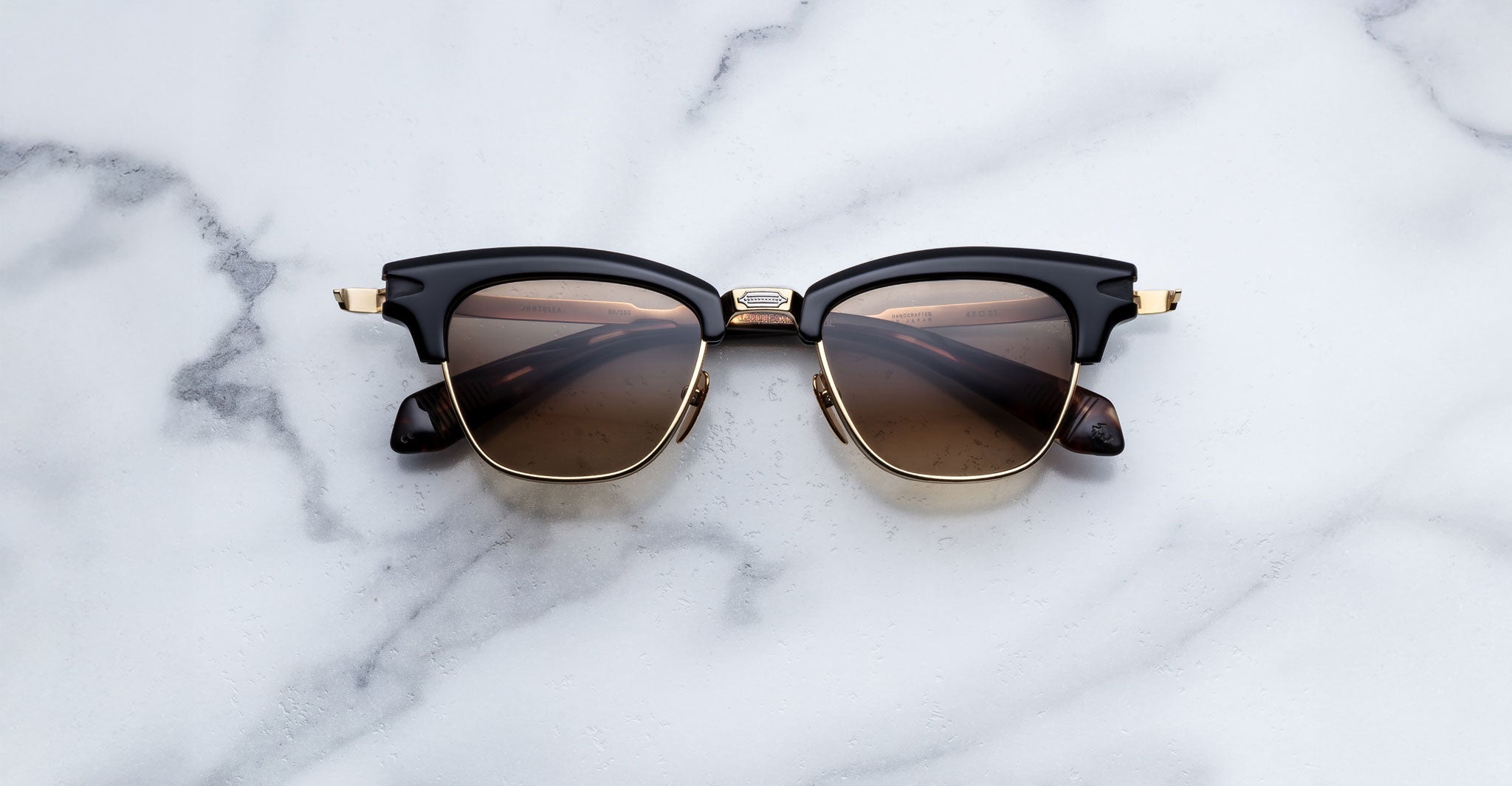 A pair of Jacques Marie Mage Sartre glasses by Jacques Marie Mage, featuring a 50s-inspired black and gold clubmaster design with dark lenses, sits on a white marble surface.