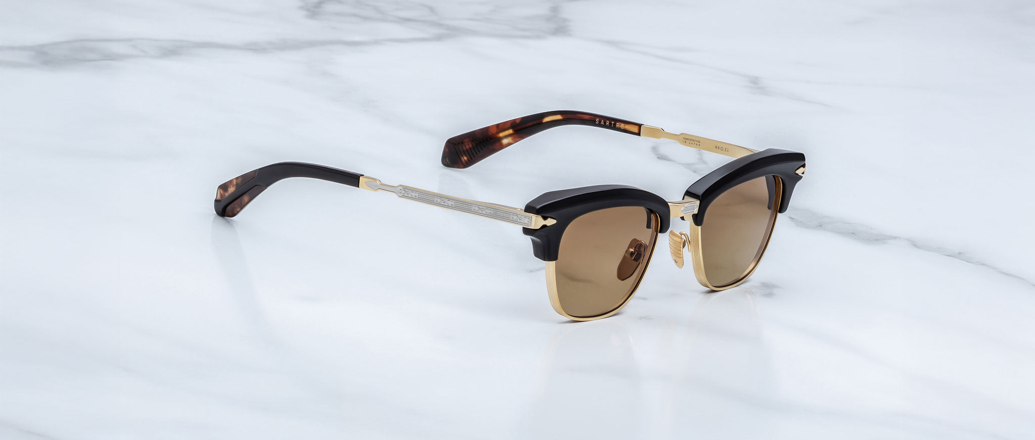 A pair of Jacques Marie Mage Sartre sunglasses with gold and black clubmaster frames and brown lenses rests on a white marble surface.