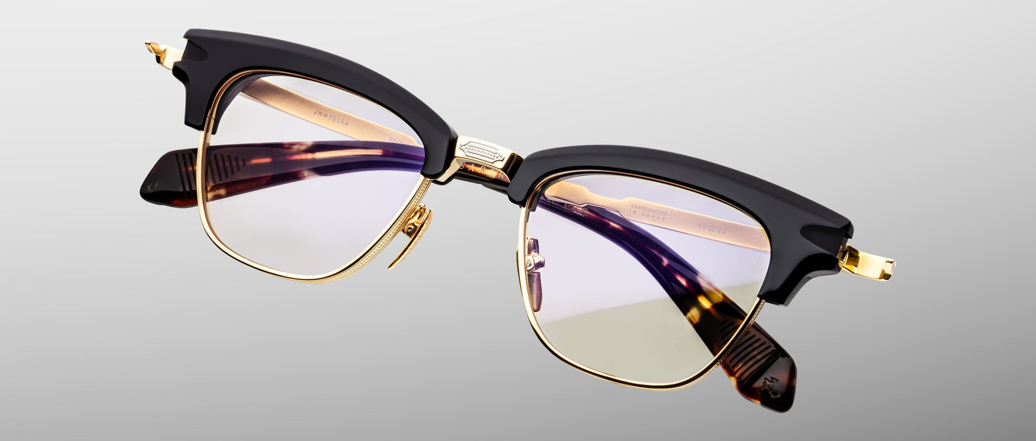 The Jacques Marie Mage Sartre by Jacques Marie Mage features 50s-inspired clubmaster style, black upper rims, a gold titanium frame, and clear lenses, displayed on a light gray background.