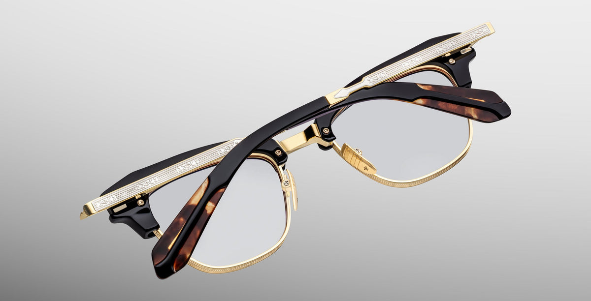 The Jacques Marie Mage Sartre glasses feature gold detailing, tortoiseshell-patterned arms, and clear lenses in a classic clubmaster frame by Jacques Marie Mage, shown against a light gray background.