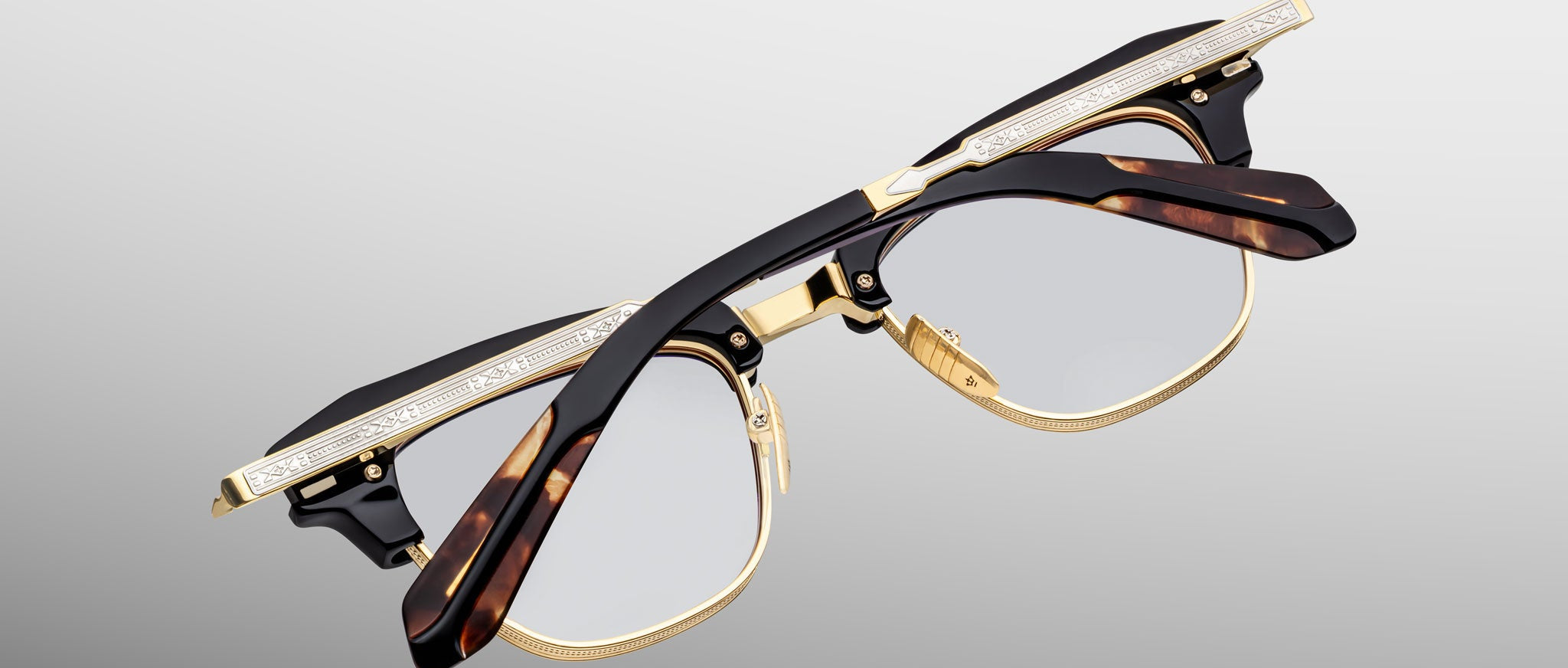 The Jacques Marie Mage Sartre glasses feature gold detailing, tortoiseshell-patterned arms, and clear lenses in a classic clubmaster frame by Jacques Marie Mage, shown against a light gray background.