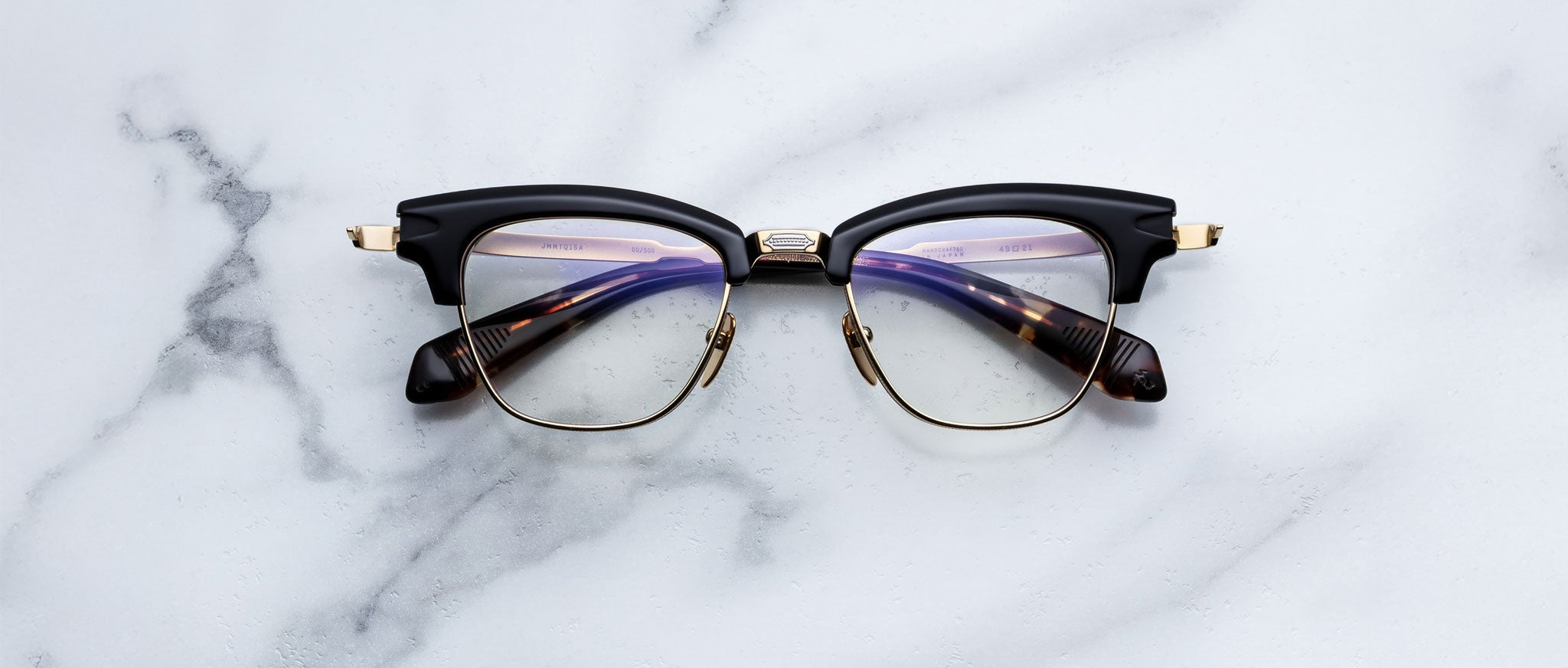 The Jacques Marie Mage Sartre by Jacques Marie Mage are 50s-inspired clubmaster glasses featuring black and gold frames with tortoiseshell arms, styled like a titanium optical frame and resting on a white marble surface.
