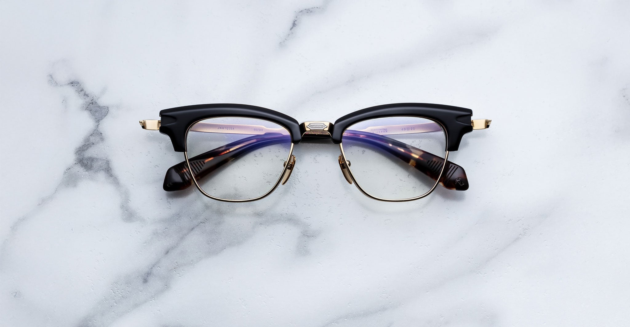 The Jacques Marie Mage Sartre by Jacques Marie Mage are 50s-inspired clubmaster glasses featuring black and gold frames with tortoiseshell arms, styled like a titanium optical frame and resting on a white marble surface.