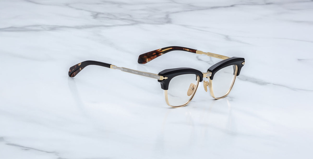 A pair of Jacques Marie Mage Sartre glasses, featuring a black and gold clubmaster frame with tortoiseshell-patterned temple tips, displayed on a white marble surface.