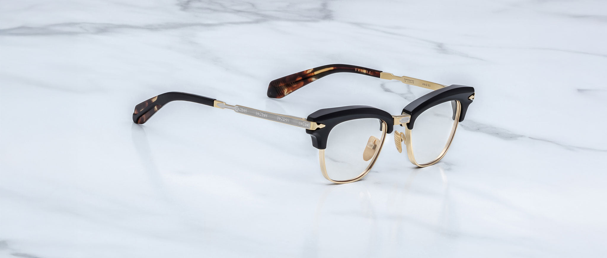 A pair of Jacques Marie Mage Sartre glasses, featuring a black and gold clubmaster frame with tortoiseshell-patterned temple tips, displayed on a white marble surface.