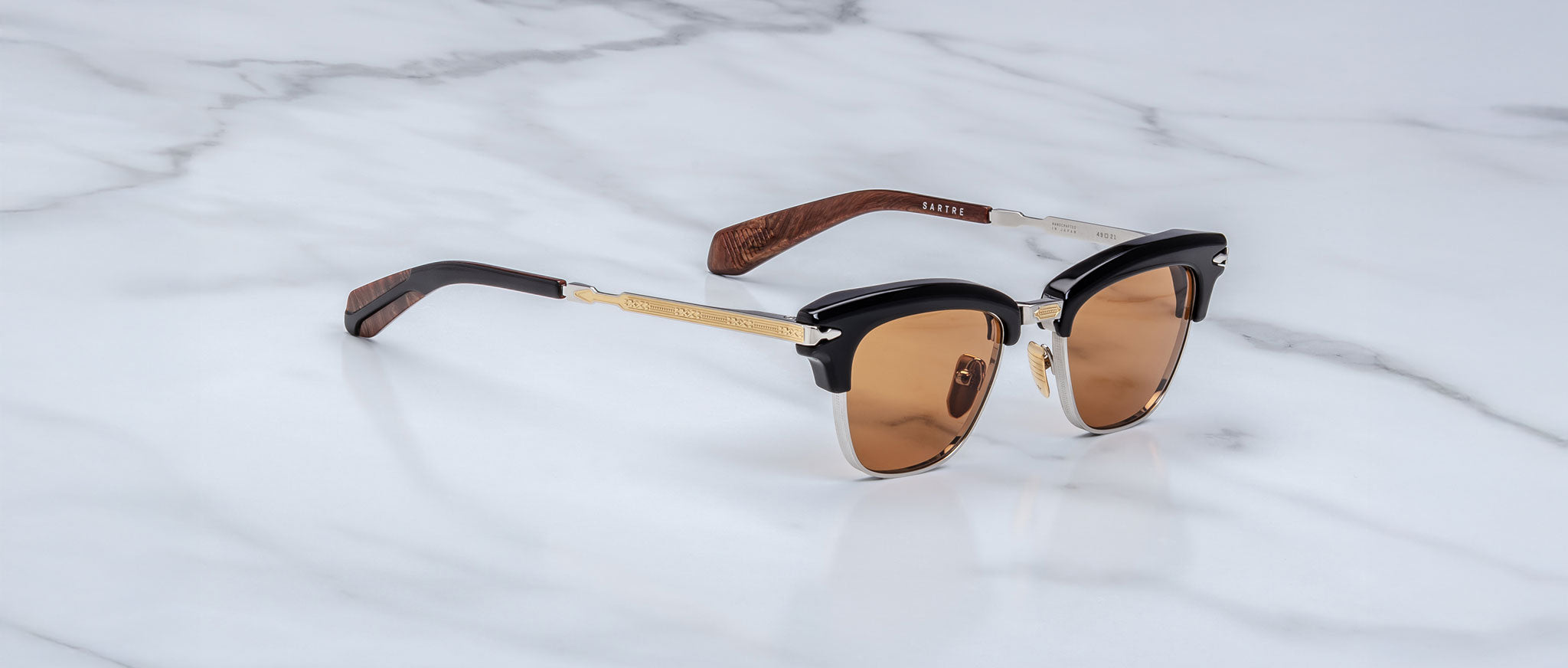 The Jacques Marie Mage Sartre sunglasses by Jacques Marie Mage feature clubmaster frames with brown-tinted lenses, black and gold accents, and wooden arms, elegantly displayed on a white marble surface.