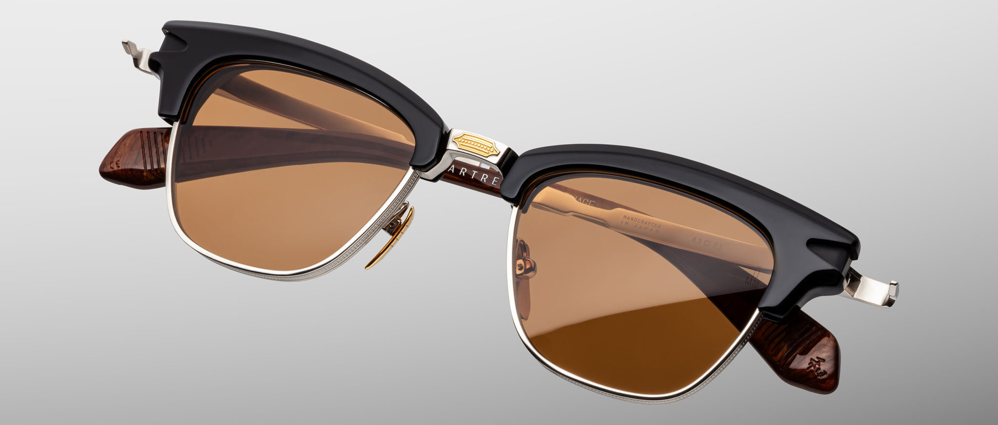 The Jacques Marie Mage Sartre sunglasses by Jacques Marie Mage feature black browline frames, gold accents, and brown tinted lenses, inspired by Sartre glasses and displayed against a plain light background.
