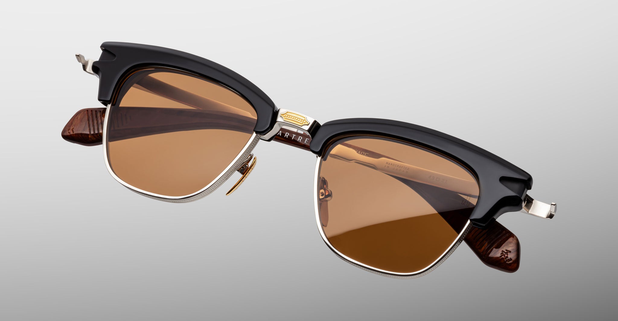 The Jacques Marie Mage Sartre sunglasses by Jacques Marie Mage feature black browline frames, gold accents, and brown tinted lenses, inspired by Sartre glasses and displayed against a plain light background.