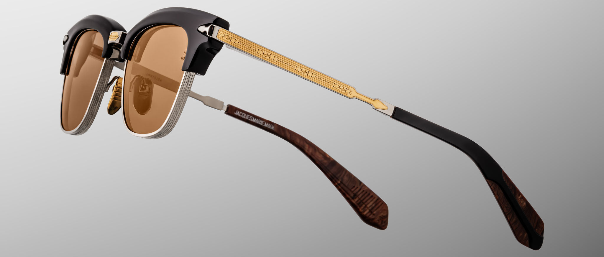 The Jacques Marie Mage Sartre sunglasses by Jacques Marie Mage feature brown lenses, black clubmaster-style upper frames, gold textured temples, and brown-patterned earpieces, shown on a gray background.