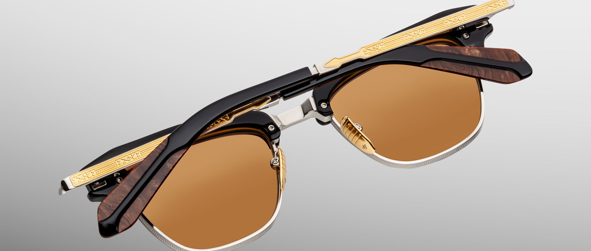 A pair of Jacques Marie Mage Sartre glasses with gold and black frames, detailed temples, and brown-tinted lenses, displayed folded on a light gray background.