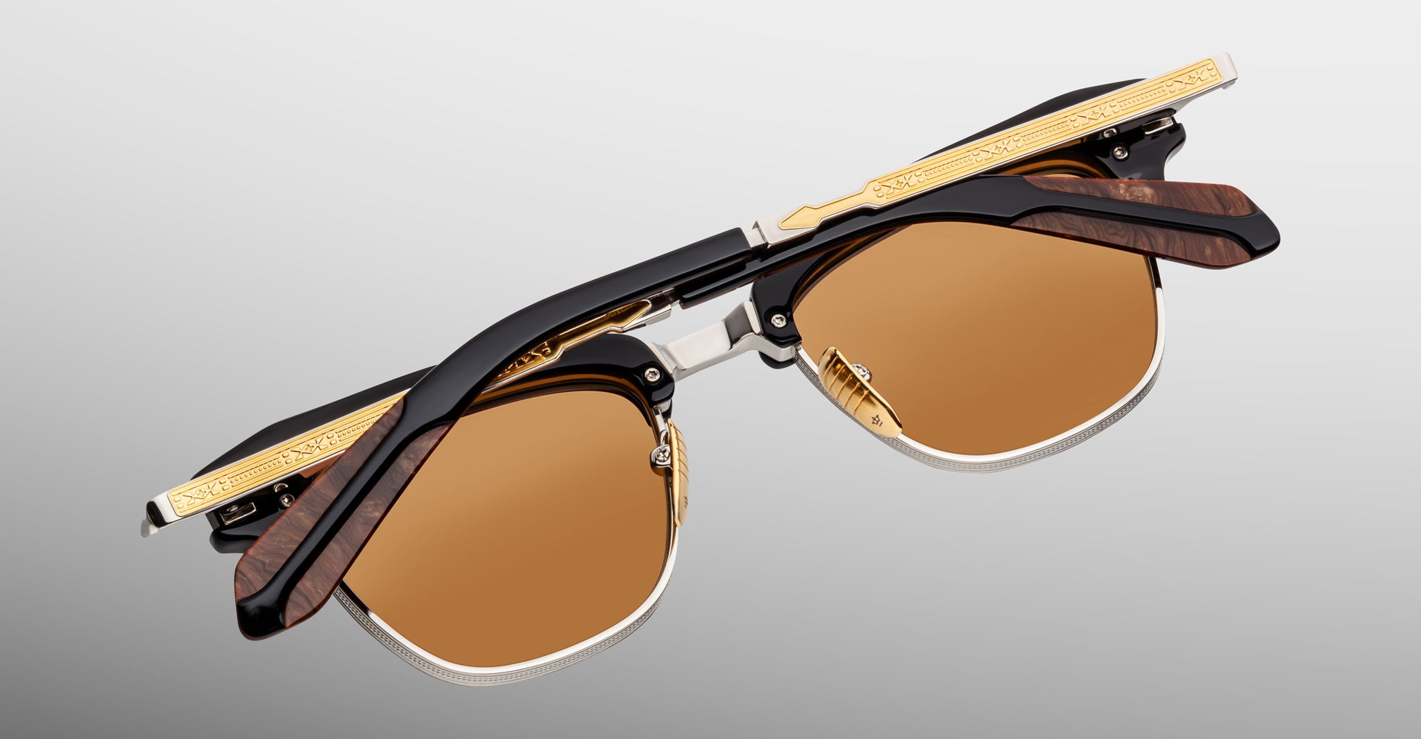 A pair of Jacques Marie Mage Sartre glasses with gold and black frames, detailed temples, and brown-tinted lenses, displayed folded on a light gray background.