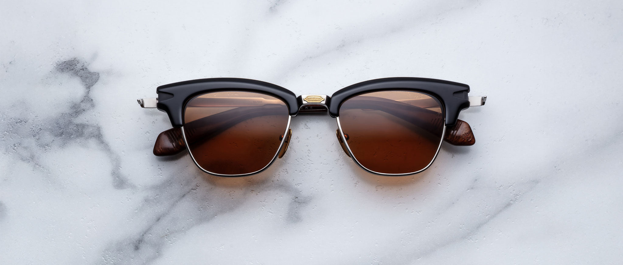 The Jacques Marie Mage Sartre glasses by Jacques Marie Mage, in black and brown with gold accents, rest elegantly on a white marble surface.