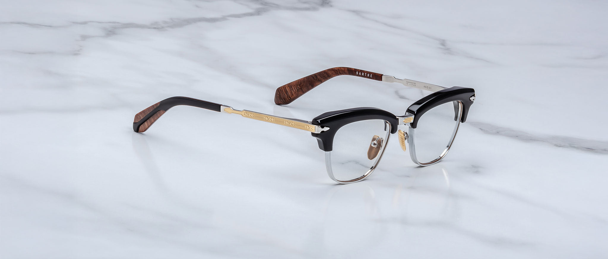 The Jacques Marie Mage Sartre glasses feature a clubmaster optical frame with black and gold accents and brown temple tips, shown on a white marble surface.