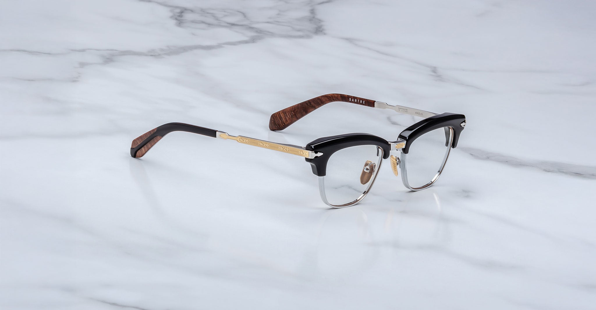 The Jacques Marie Mage Sartre glasses feature a clubmaster optical frame with black and gold accents and brown temple tips, shown on a white marble surface.