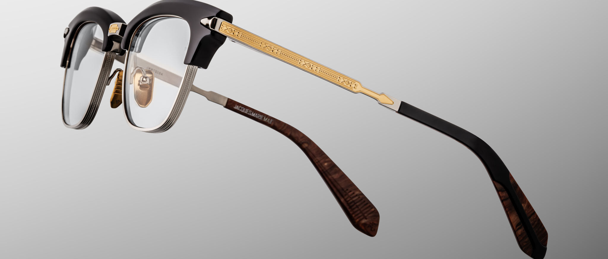 The Jacques Marie Mage Sartre glasses by Jacques Marie Mage feature a black and silver frame, gold decorative temples, clear lenses, and brown patterned temple tips, elegantly showcased on a gray gradient background.