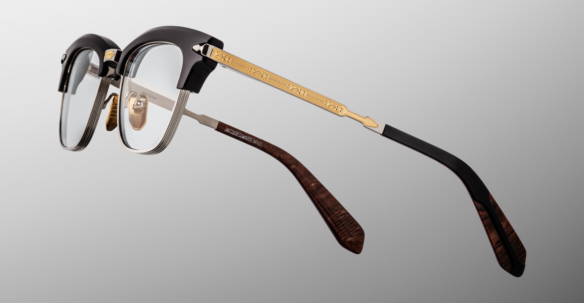 The Jacques Marie Mage Sartre glasses by Jacques Marie Mage feature a black and silver frame, gold decorative temples, clear lenses, and brown patterned temple tips, elegantly showcased on a gray gradient background.