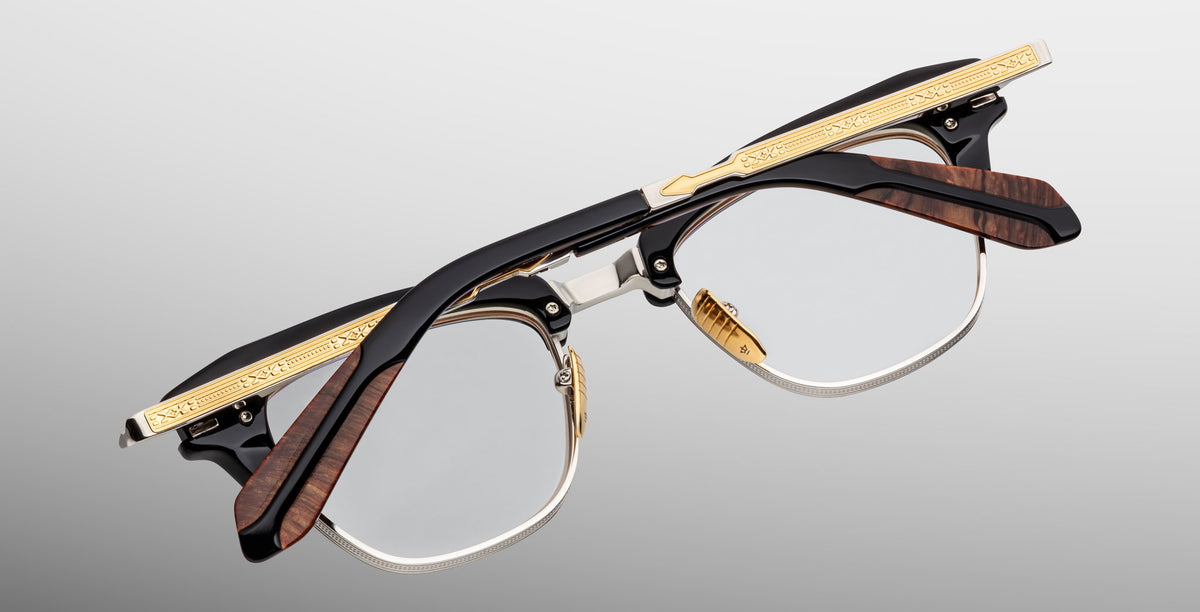 A pair of Jacques Marie Mage Sartre glasses featuring black, gold, and brown wood-textured clubmaster frames is shown upside down against a gray gradient background.