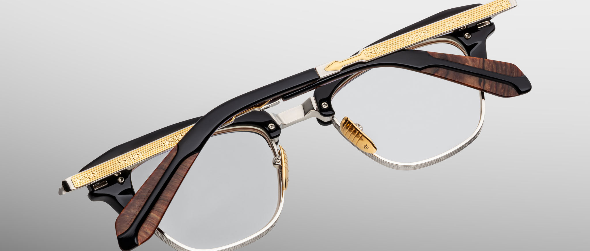 A pair of Jacques Marie Mage Sartre glasses featuring black, gold, and brown wood-textured clubmaster frames is shown upside down against a gray gradient background.